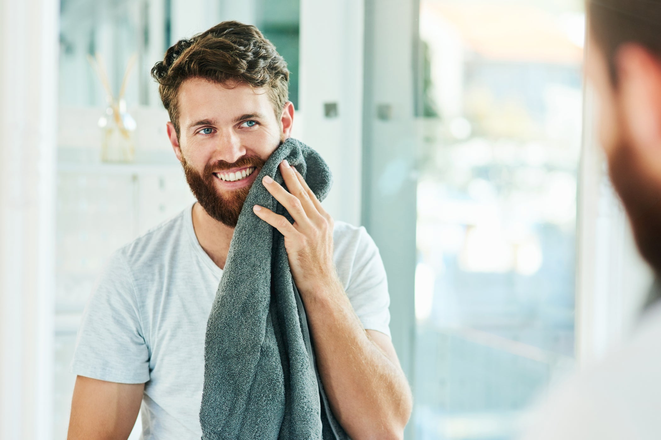 Junger Mann mit braunen Haaren und Bart steht im Bad und tupft sich das Gesicht mit einem Handtuch ab