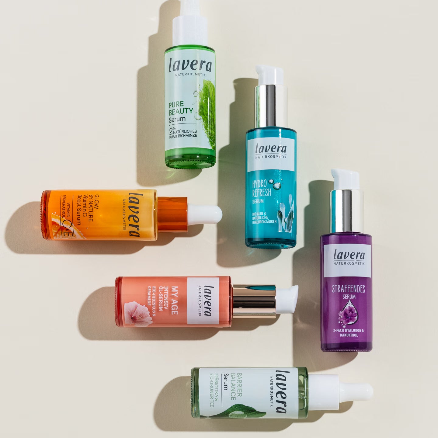 Six lavera facial serums in differently colored bottles are lying and standing on a light-colored surface in the sunlight.