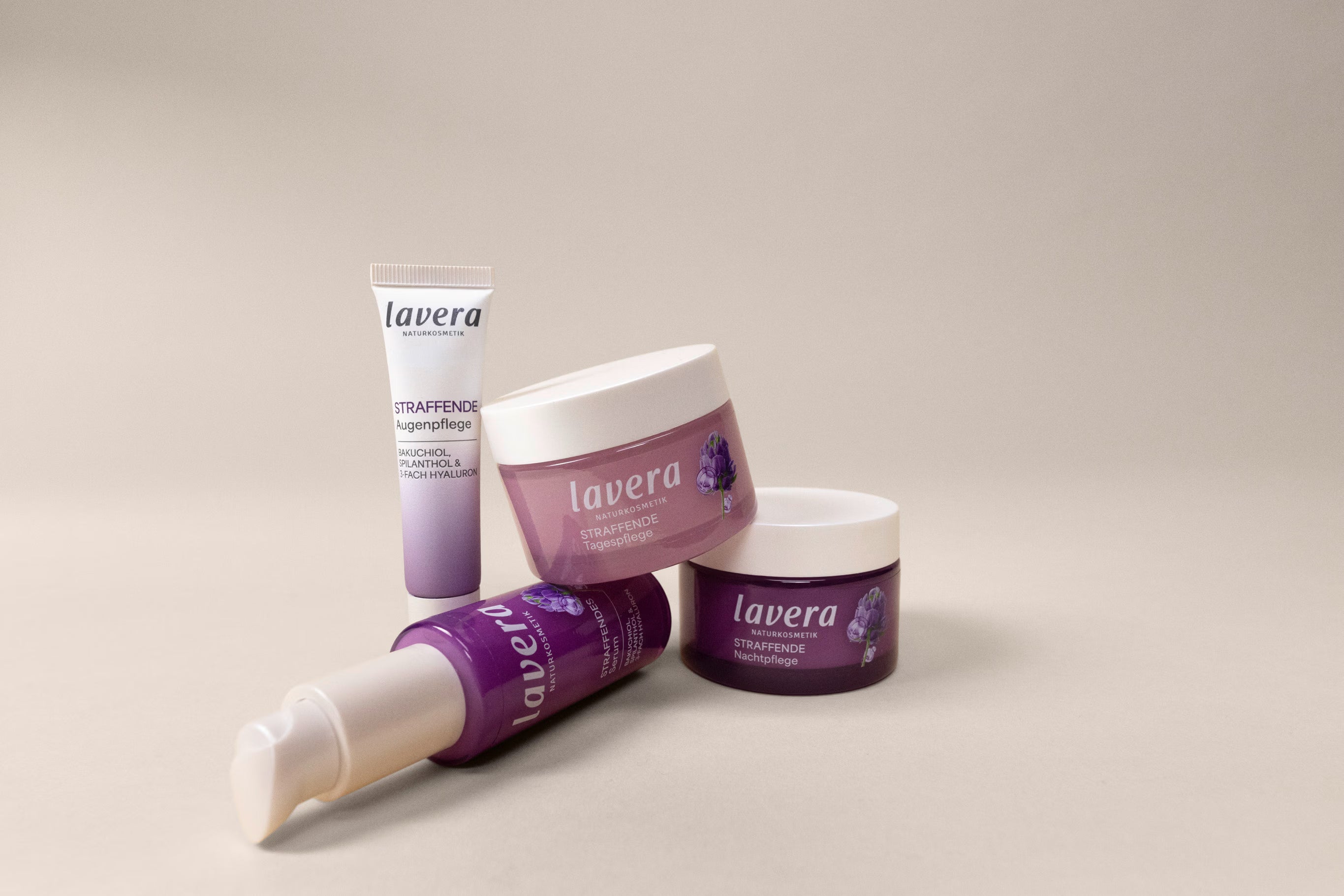 Four lavera firming facial care products in purple packaging are lying and standing against a light background.