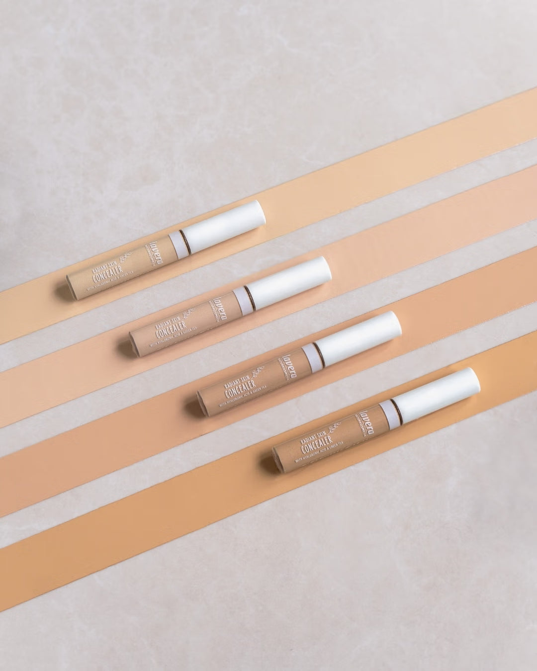 Four lavera Radiant Skin Concealers in different shades on a beige striped pattern.