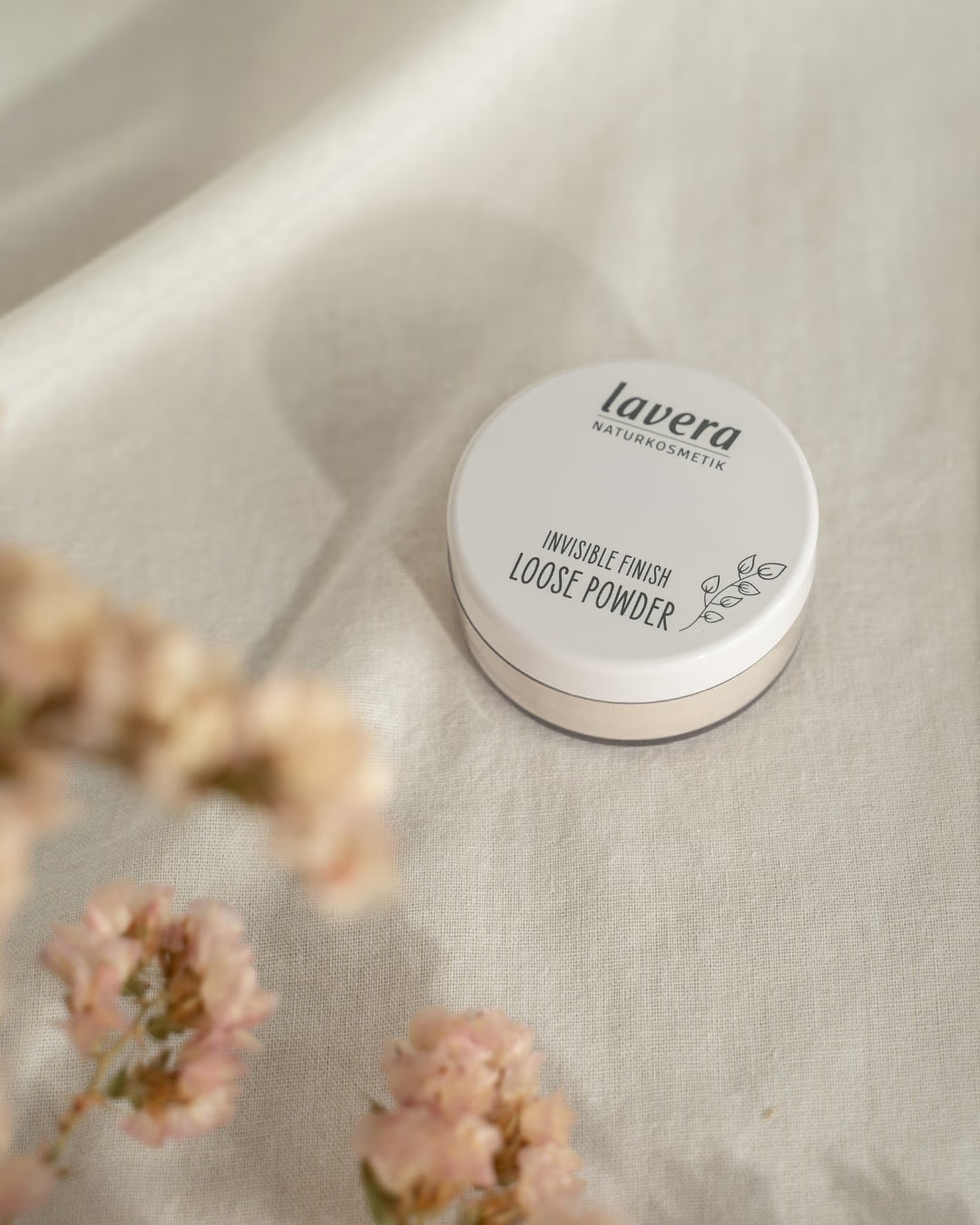 lavera Invisible Finish Loose Powder lies on light linen fabric, surrounded by blurred pink blossoms.