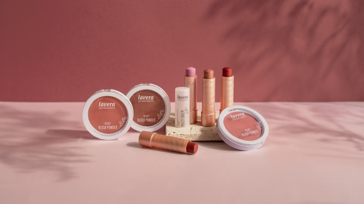 Several lavera blush products and multi-balms in rose, nude, and red shades are displayed on a pedestal against a pink background.