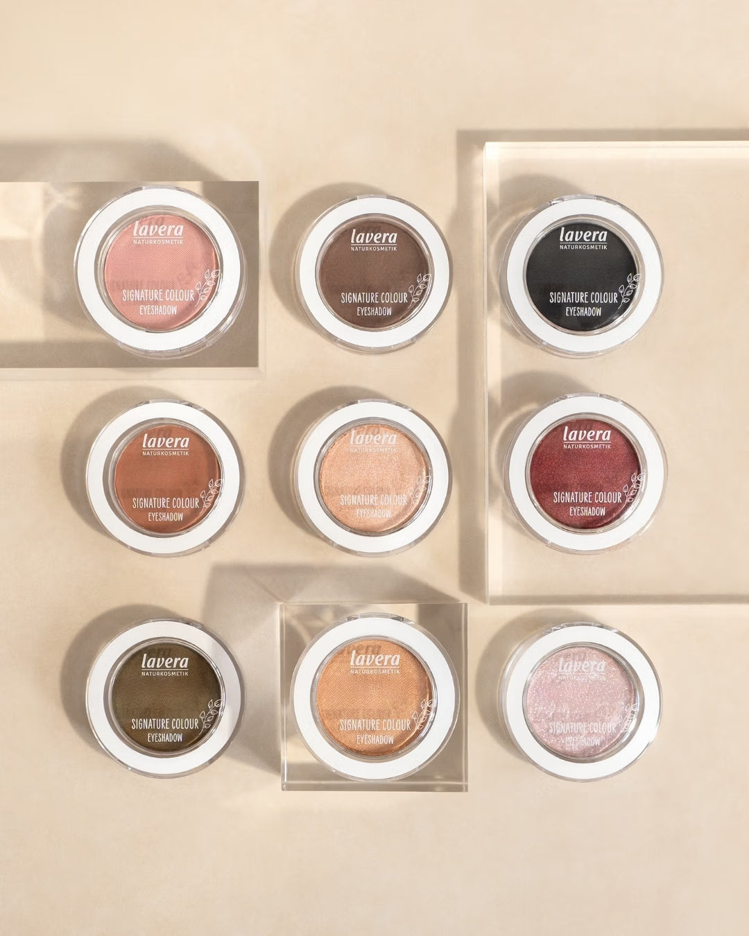 Nine lavera Signature Colour eyeshadows in various shades are displayed on transparent acrylic blocks against a beige background.