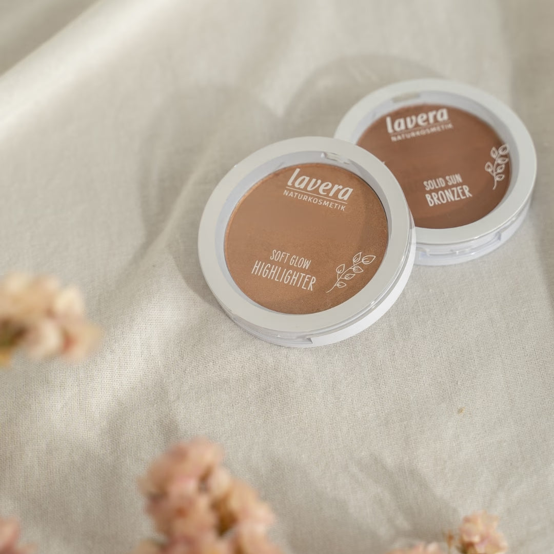 lavera complexion products, a Soft Glow Highlighter and a Solid Sun Bronzer, lie on light linen fabric beside blurred pink flowers.