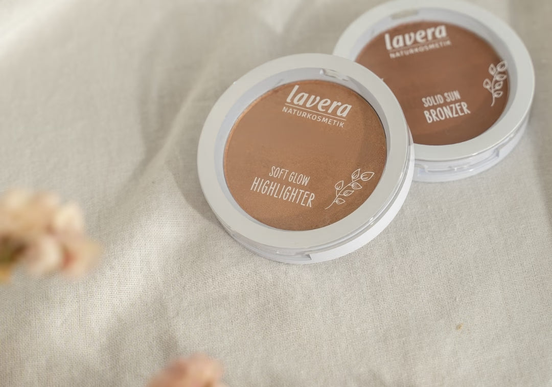 lavera complexion products, a Soft Glow Highlighter and a Solid Sun Bronzer, lie on light linen fabric beside blurred pink flowers.