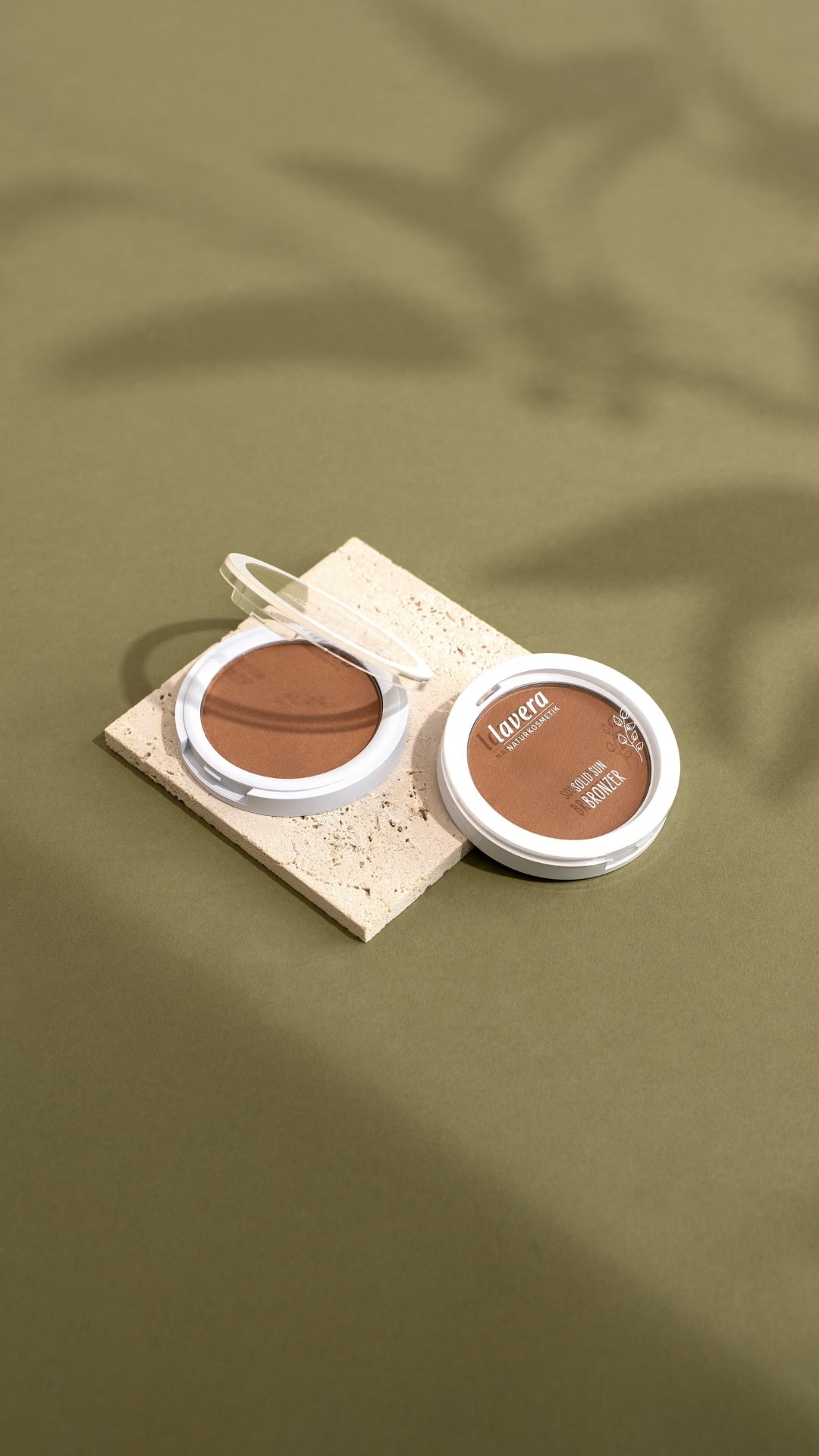 Two opened lavera complexion products - highlighter and Solid Sun Bronzer - are placed on a light stone pedestal against an olive-green background.