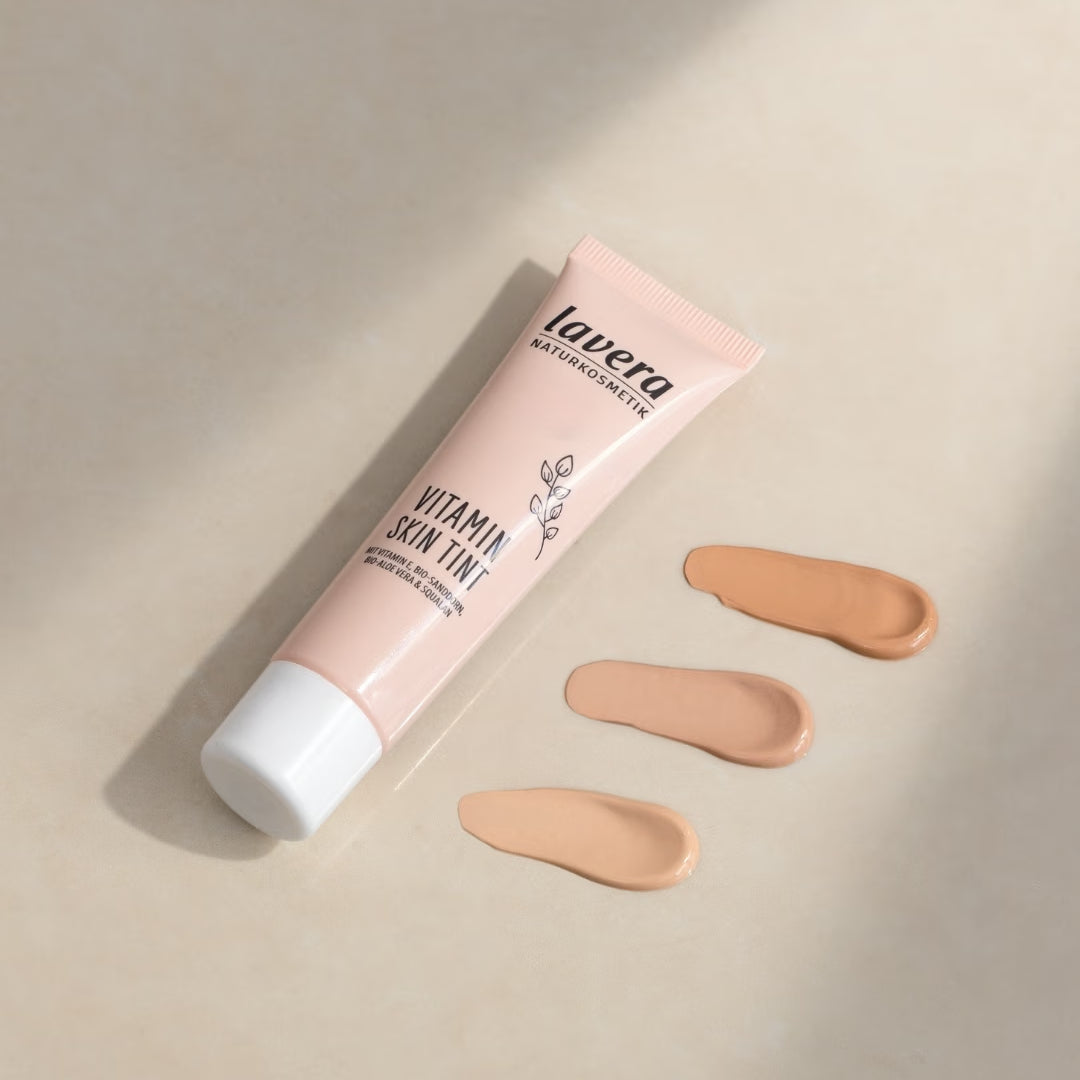 A tube of lavera Vitamin Skin Tint lies next to three swatches of varying shades on a beige surface.