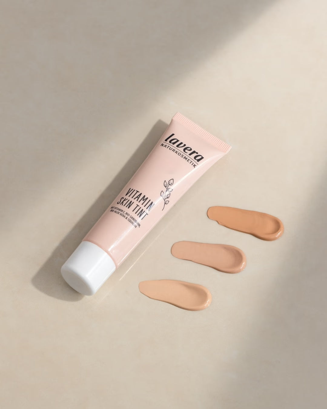 A tube of lavera Vitamin Skin Tint lies next to three swatches of varying shades on a beige surface.