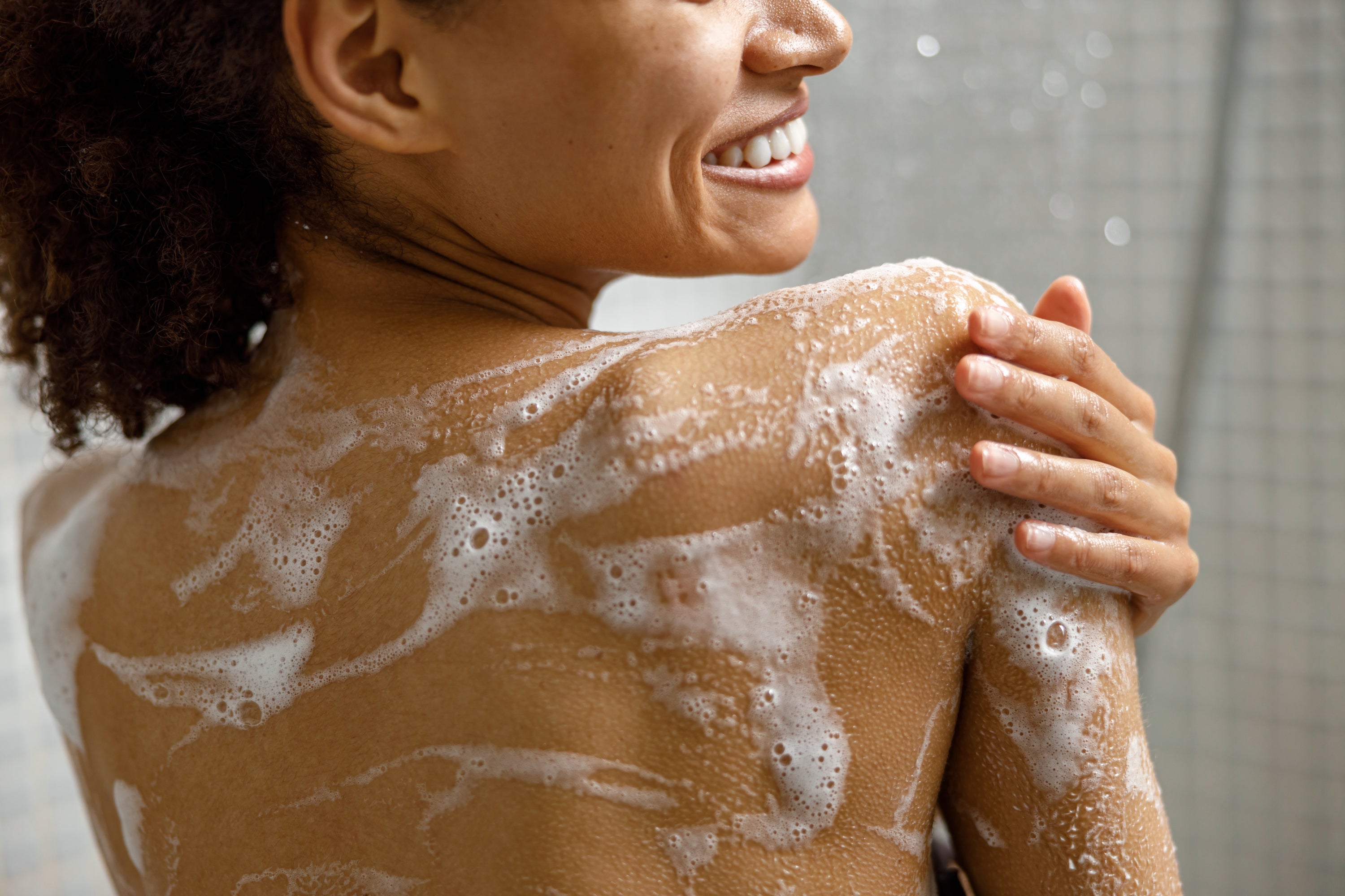 Nahaufnahme des oberen Rückens einer weiblichen Person unter der Dusche mit Duschgel auf der Haut