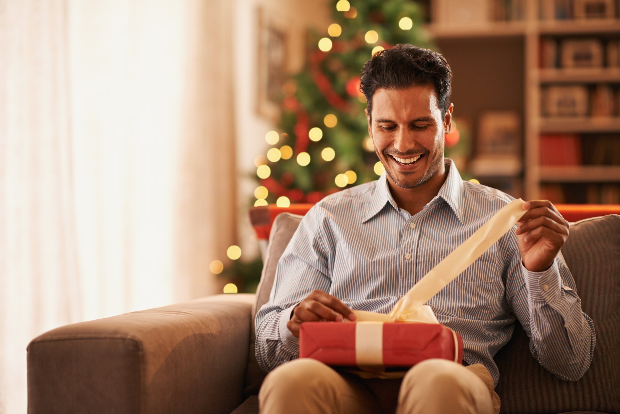 Mann sitzt auf einem Sofa und packt ein Geschenk aus – mit Weihnachtsbaum im Hintergrund