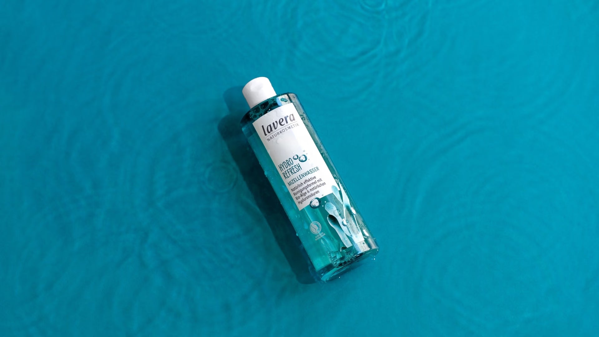 A bottle of lavera Hydro Refresh micellar water lies on a brightly turquoise-blue, textured surface in the sunlight.