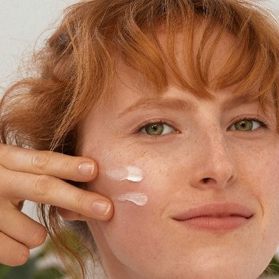 Rothaarige Frau trägt Creme auf die Wange auf, zwei Punkte sichtbar