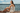 A young woman sitting sideways on a pier at a lake, facing the sun with her face