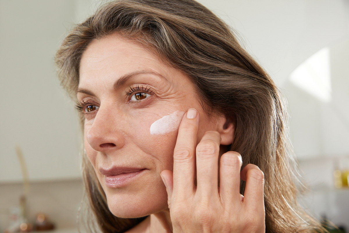 Elderly woman with long, dark blonde hair applies cream to her cheek with her finger