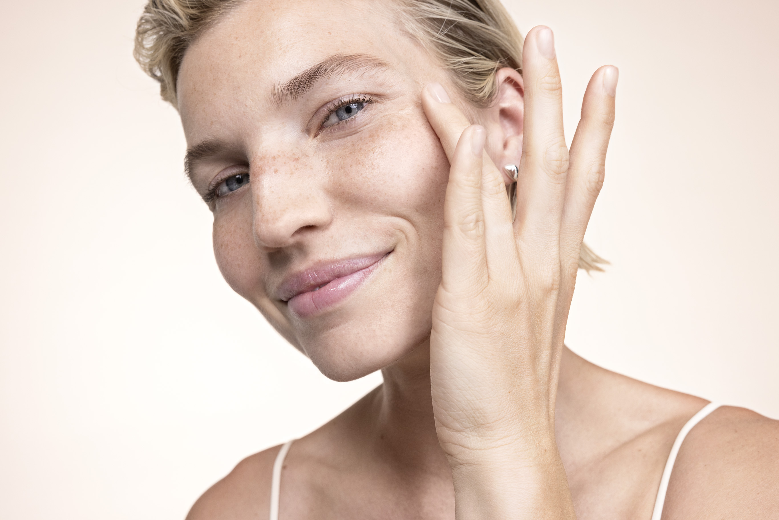 A blonde woman strokes her cheek with a finger and smiles.