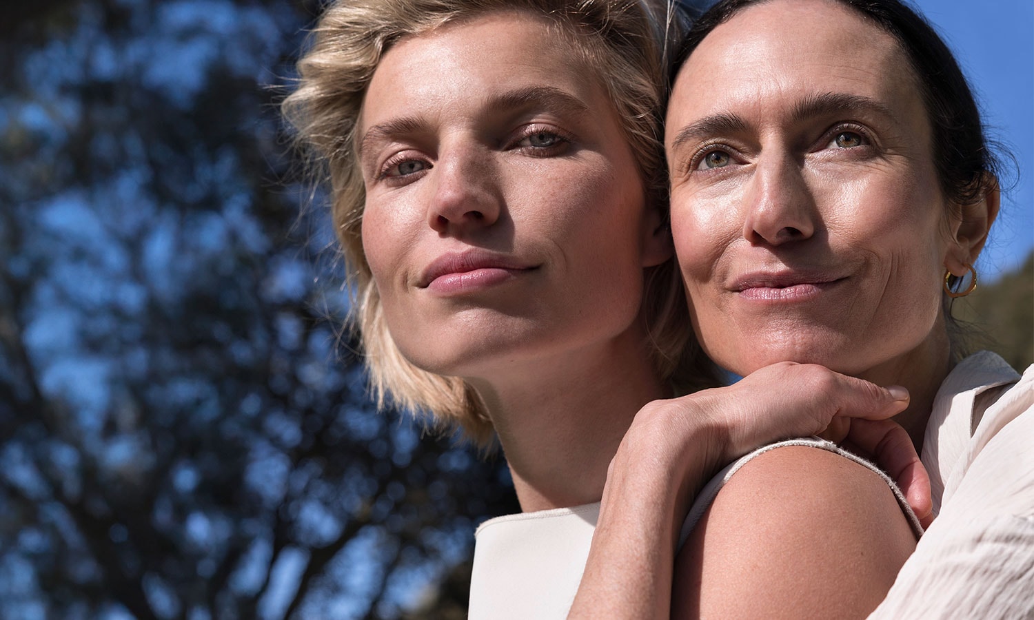 A blonde and a dark-haired model are standing in front of a tree, holding on to each other.