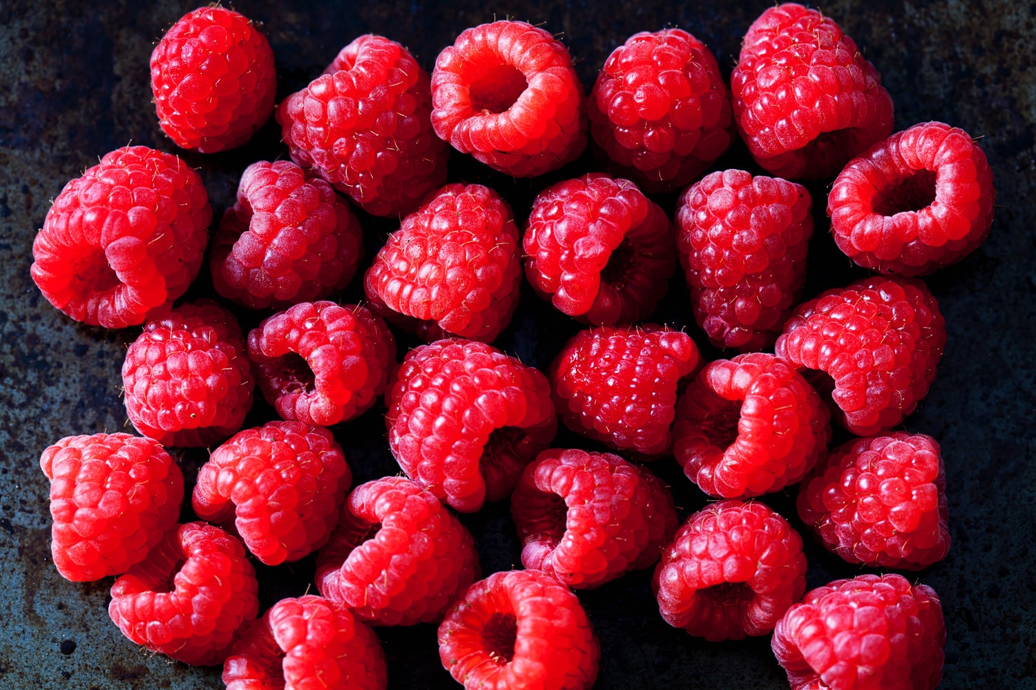 Bio-Himbeeren wirken antioxidativ Nahaufnahme von zahlreichen roten Himbeeren auf dunklem Tisch