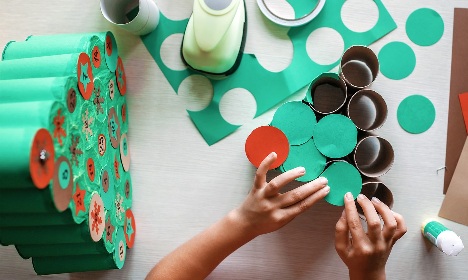 Frauenhände basteln an einem selbst aus Klopapierrollen zusammengeklebten Tannenbaum mit Bastelutensilien