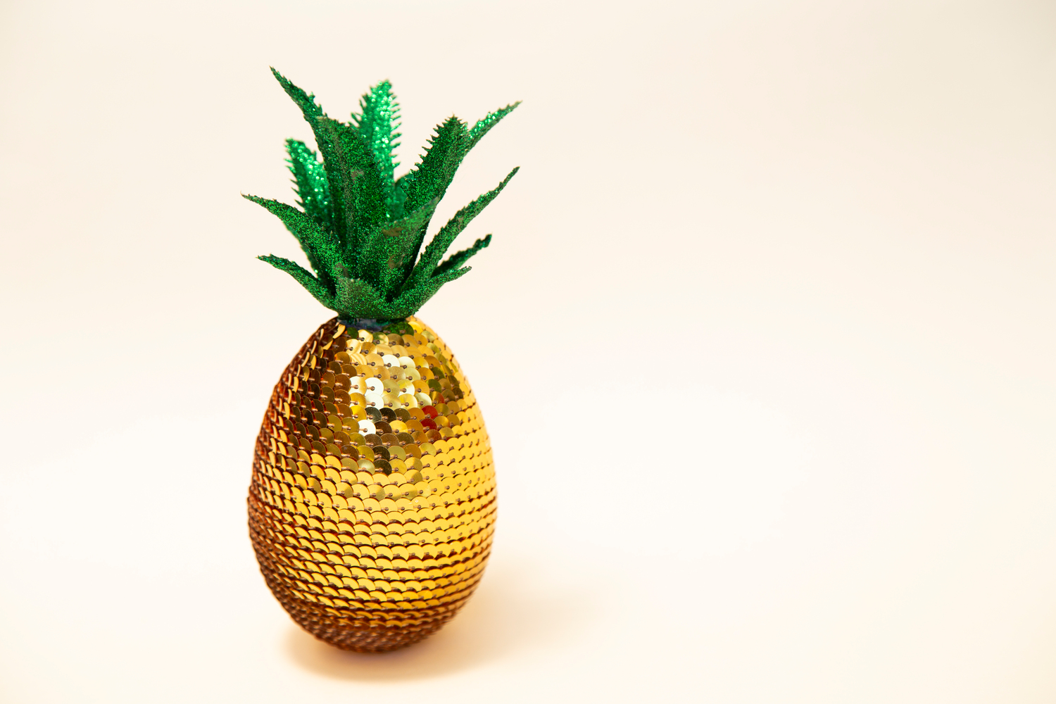 Ananas aus goldenen Pailletten und gruenen stoffblaettern vor cremefarbenen Hintergrund