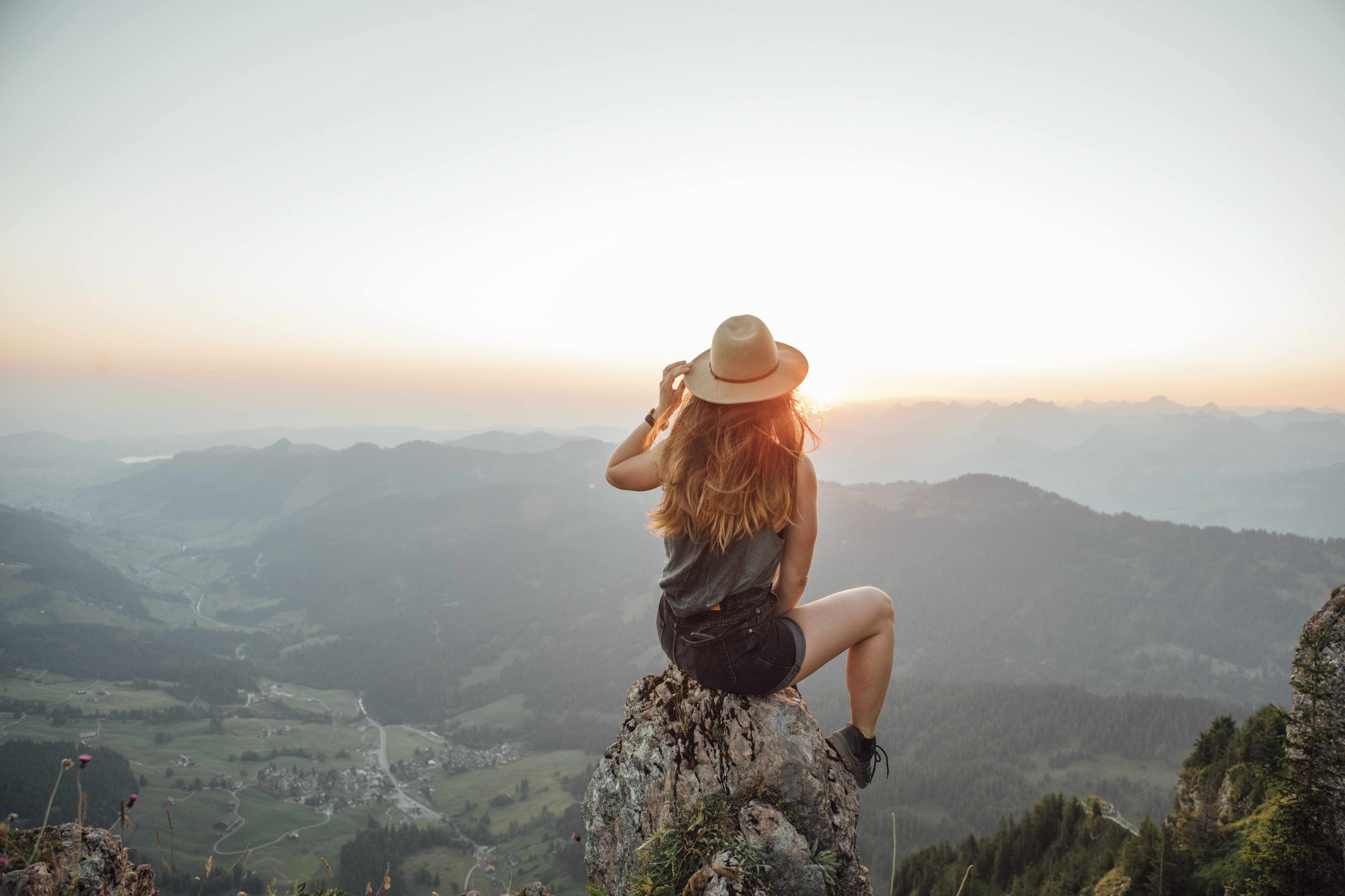 Frau mit langen braunen Haaren und Hut befindet sich auf einem Berg sitzt mit dem Rücken zur Kamera auf einem Stein und blinkt in die Ferne
