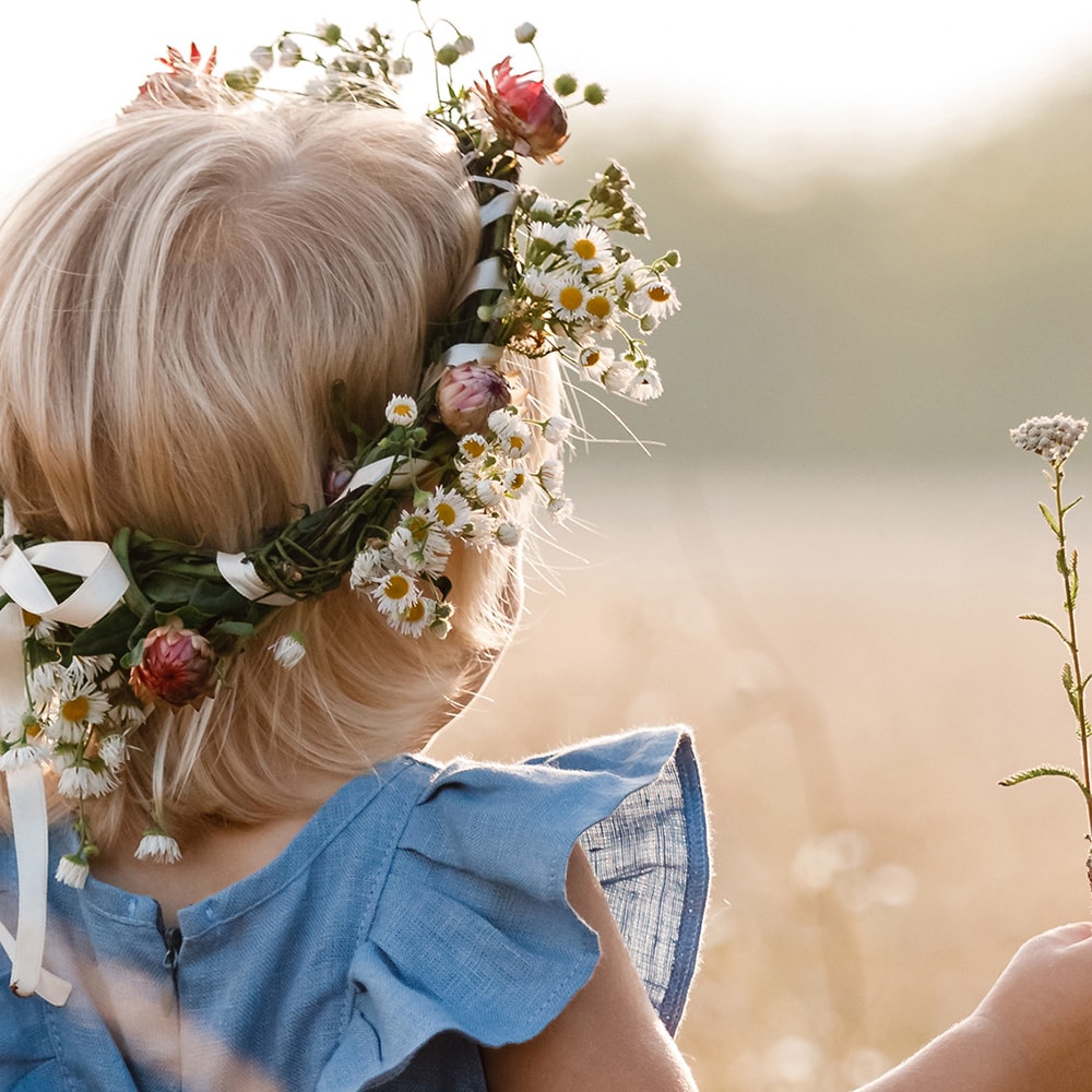 Ein kleines blondes Mädchen steht vor einem Getreidefeld und trägt ein bunten Blumenkranz im Haar