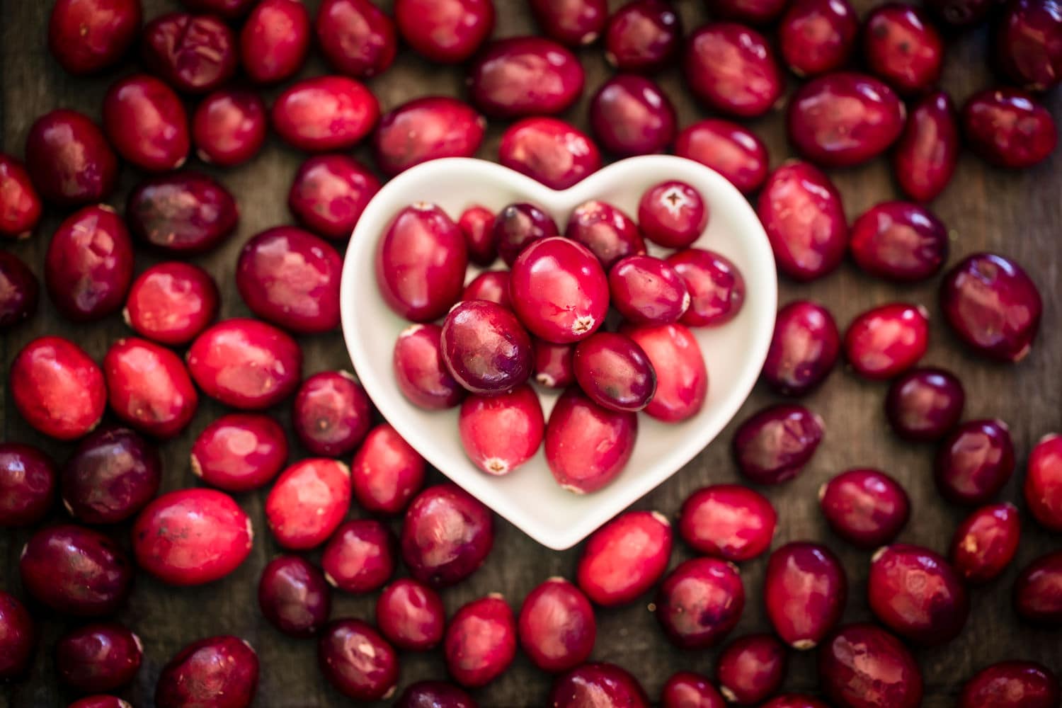Naturwunder Cranberry für deine Hautpflege Weiße Porzellanschale gefüllt mit einigen dunkelroten Cranberries und losen Cranberries auf dunklem Holztisch